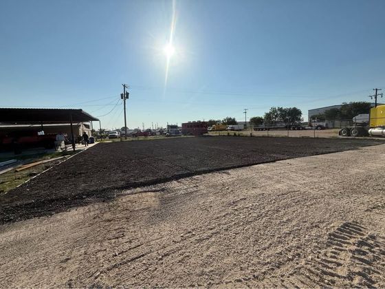 A dirt lot with dark soil is in the foreground. The sun shines brightly in the sky. Buildings are in the distance.