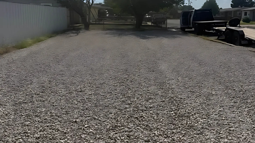 A gravel driveway leads to a gate, with a fence and parked trucks visible in the distance.