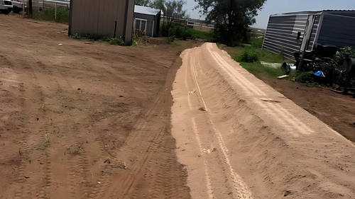 Dirt road with tire tracks, leading uphill, with buildings in the background.