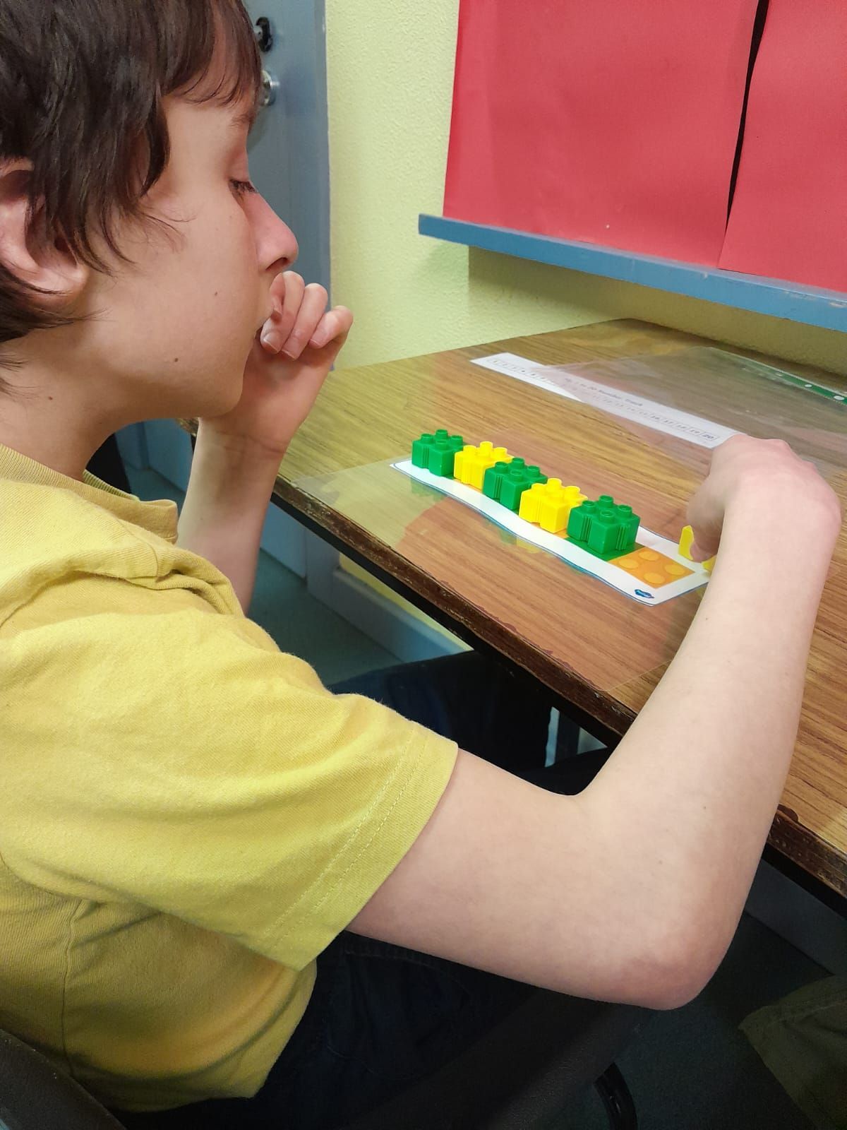 A boy in a yellow shirt sits at a desk with a ruler and blocks on it