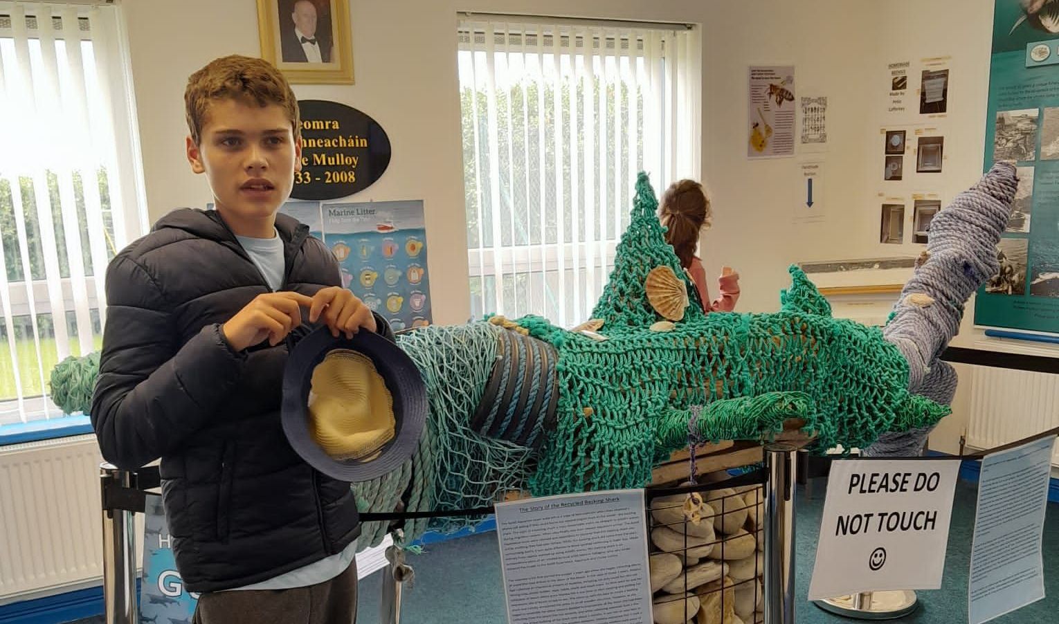 A boy is holding a hat in front of a sculpture of a shark.