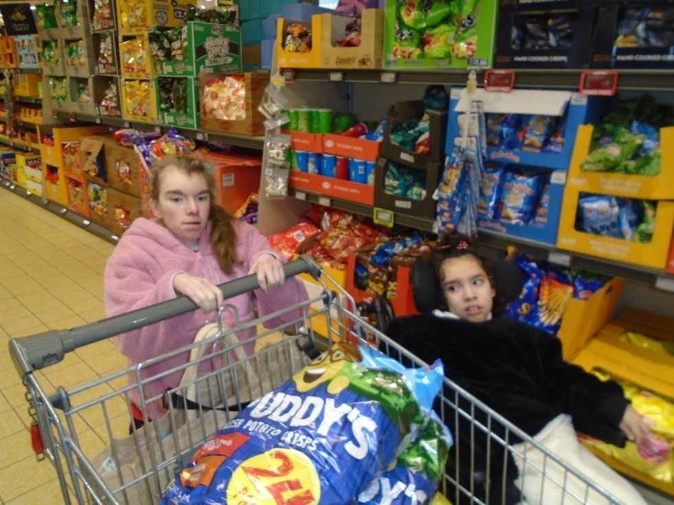 Two girls in a shopping cart with a bag of daddy 's chips