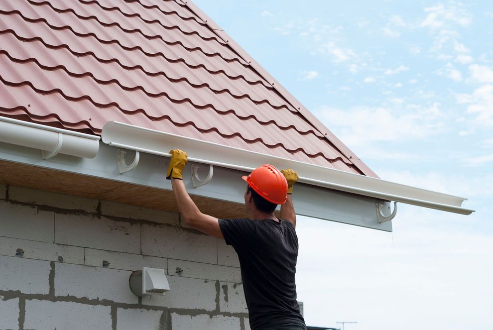 Worker in an orange hard hat cleaning gutters beneath a red roof outdoors