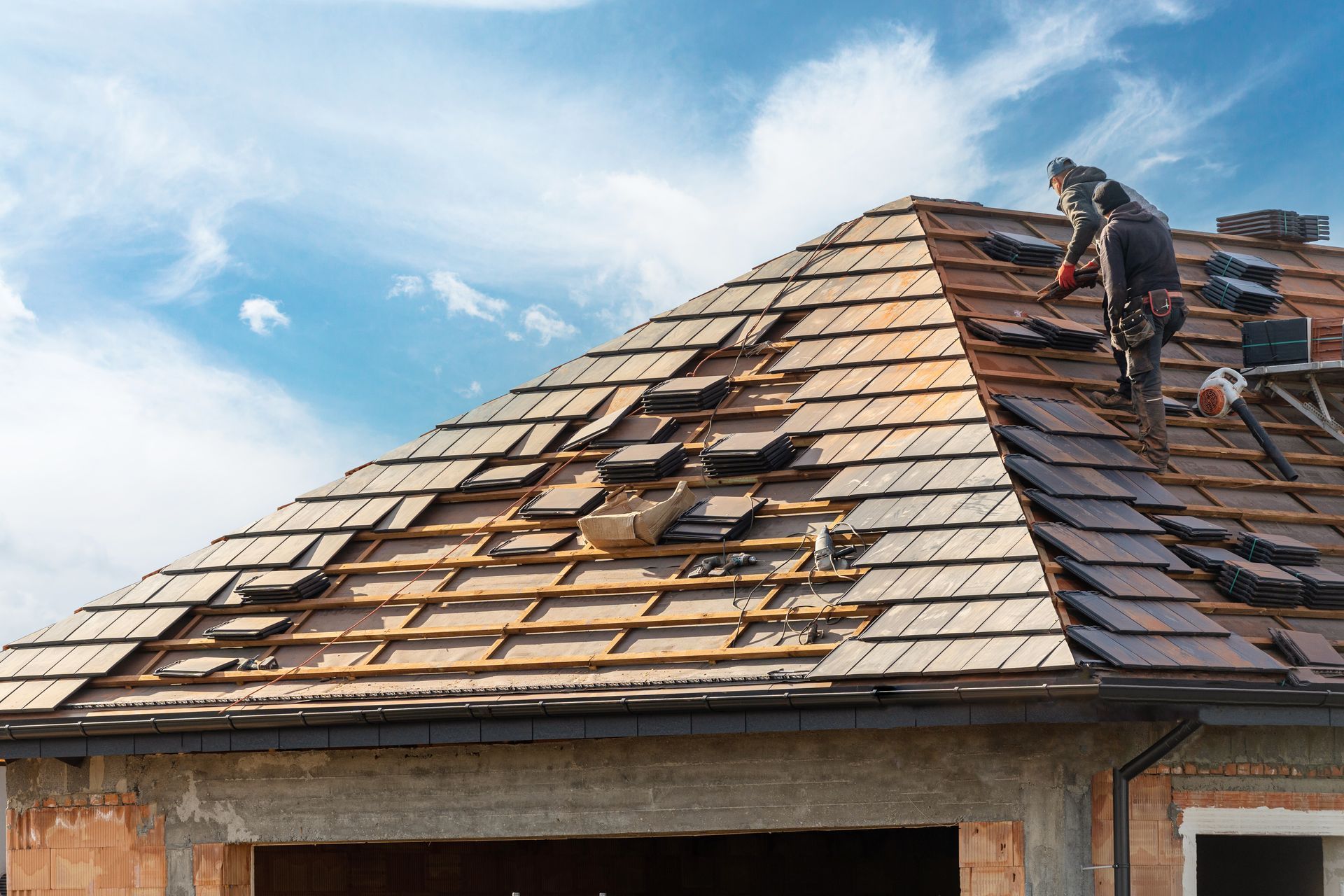 Workers repairing a shingled roof on a house under a blue sky
