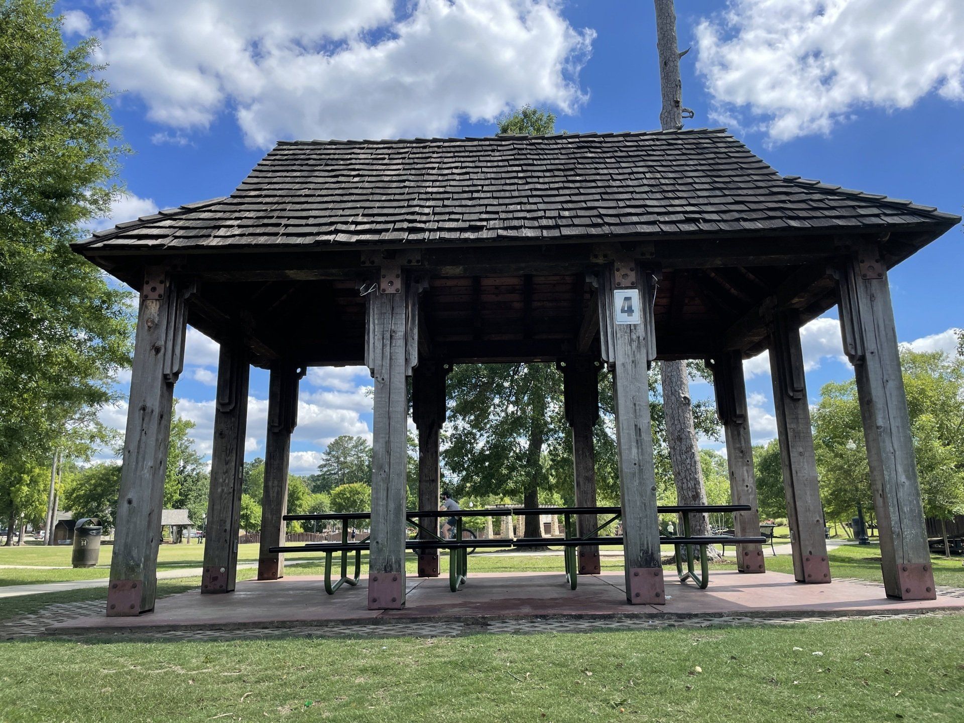 A wooden gazebo with a roof and a picnic table in a park.