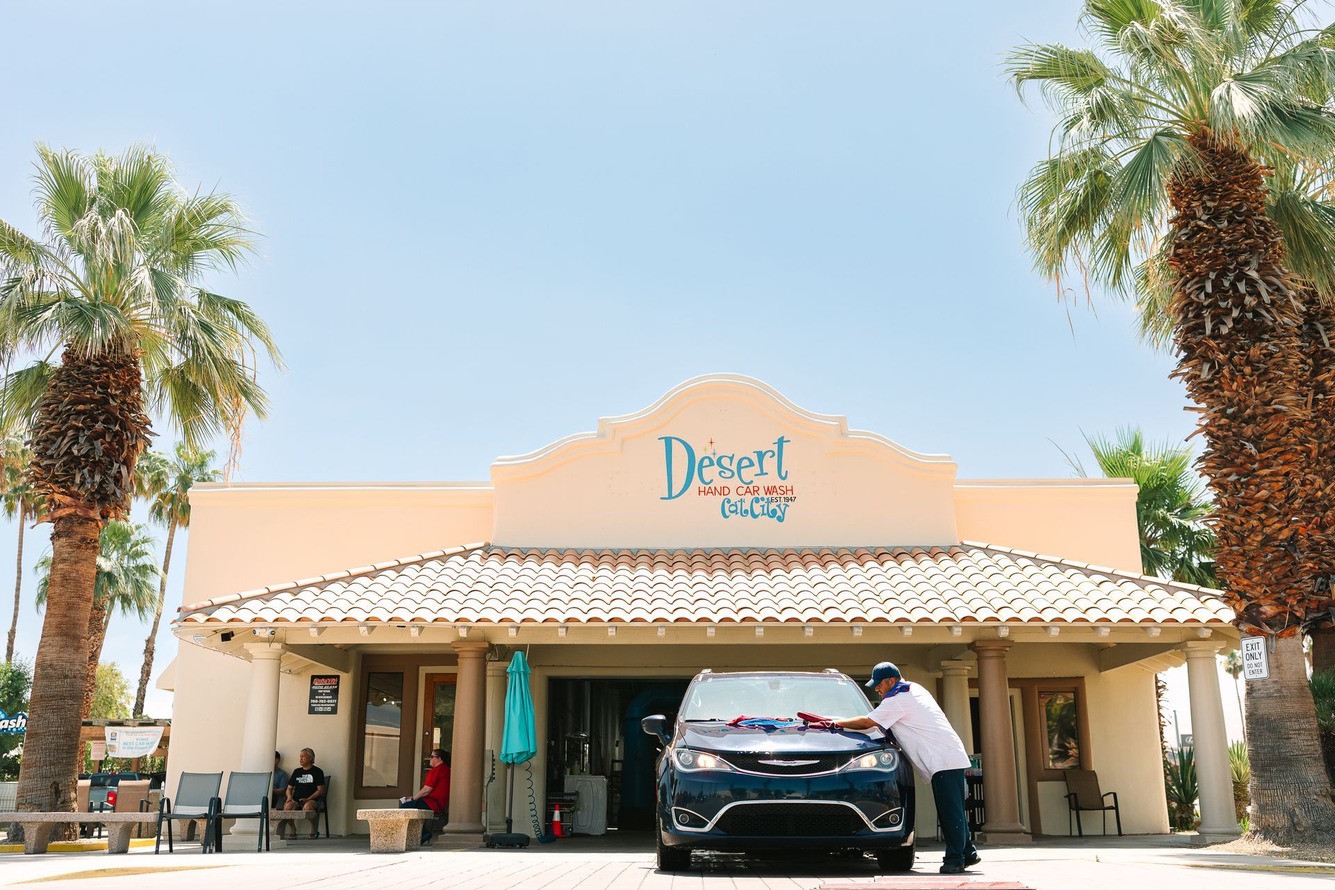 A man is washing a car in front of a building with palm trees.