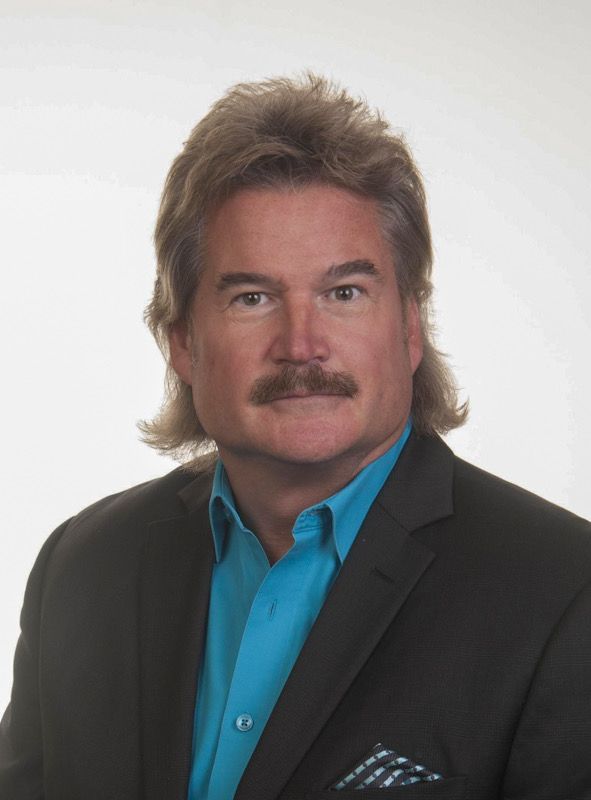 A man with long hair and a mustache is wearing a suit and a blue shirt.