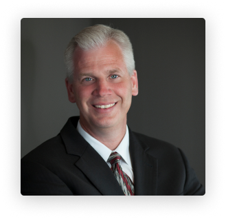 Studio headshot of a smiling man in a dark suit and striped tie against a gray background