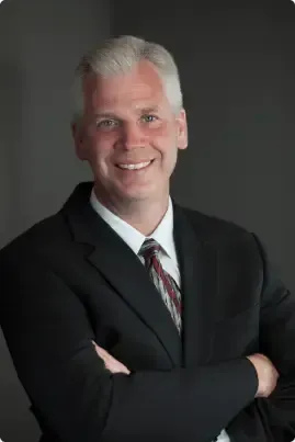 Smiling man in a black suit with arms crossed against a dark gray background