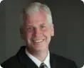 Smiling man in a dark suit and white shirt against a gray background