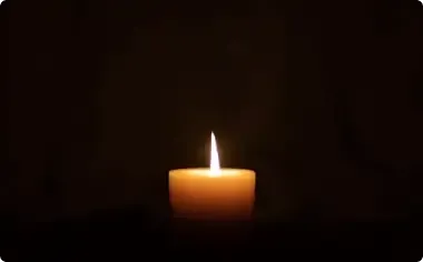 A lit candle glowing in a dark room, with a small flame and warm orange wax.