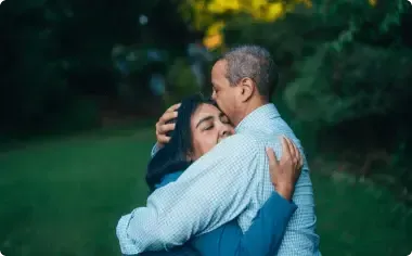 Two people embracing outdoors in a green park, one in a light blue shirt and the other in a blue vest