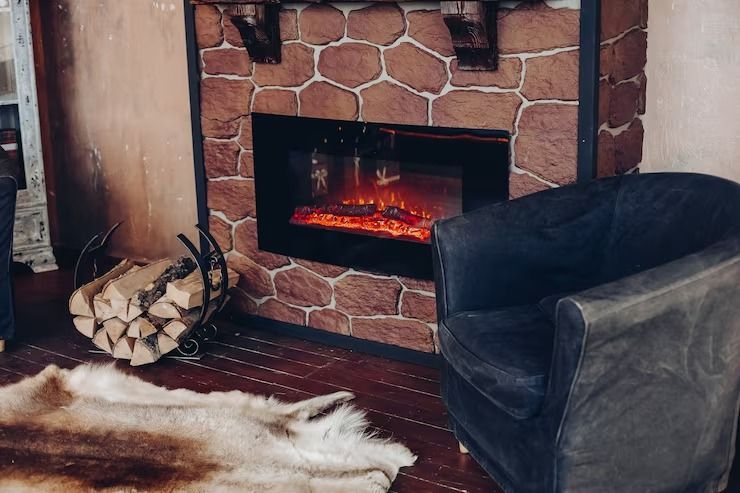 A cozy corner with a stone fireplace, a black armchair, a stack of firewood in a metal holder, and a furry rug.