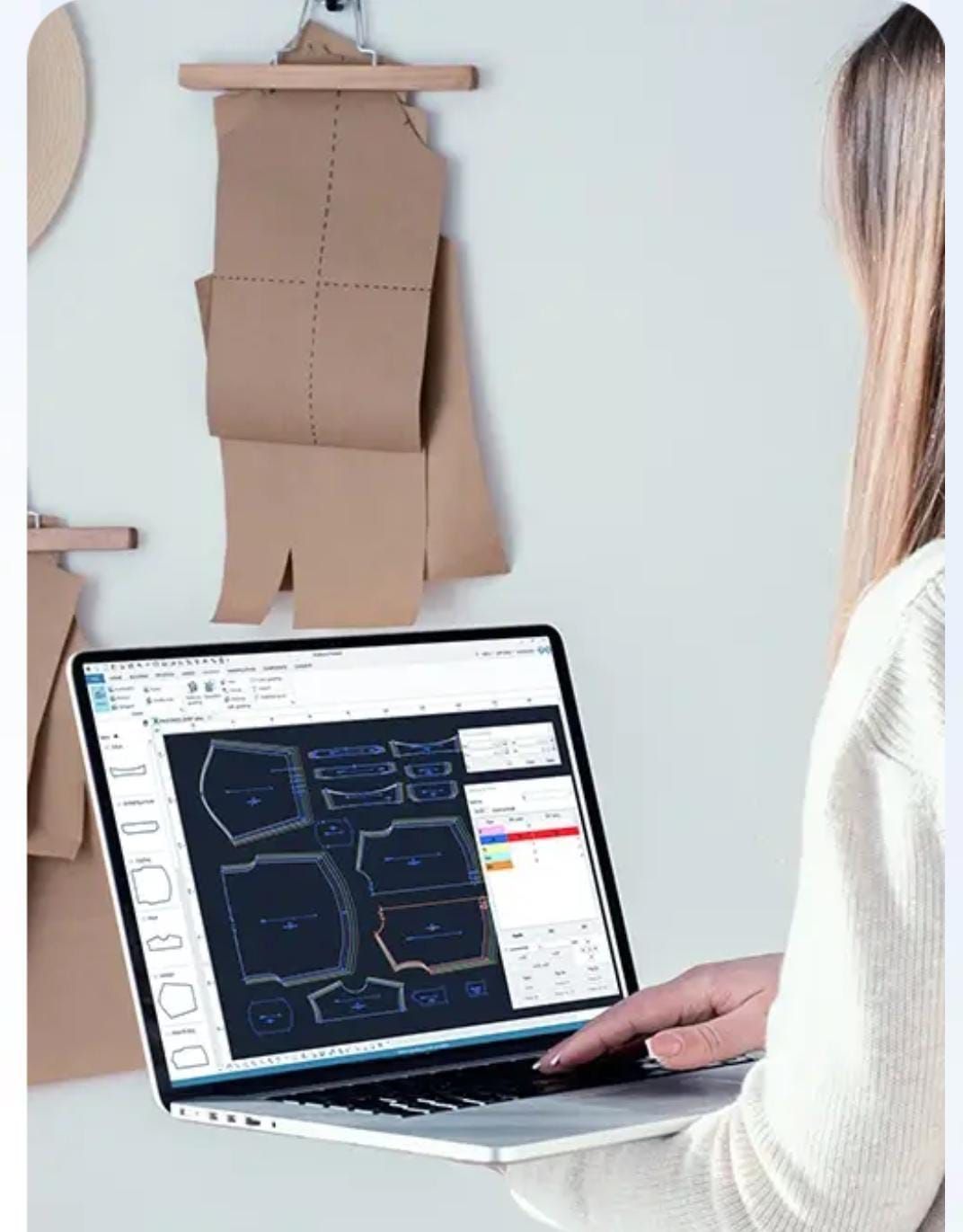 Persona utilizando un ordenador portátil mostrando un patrón de prenda junto a piezas de patrones de papel colgadas en una pared.