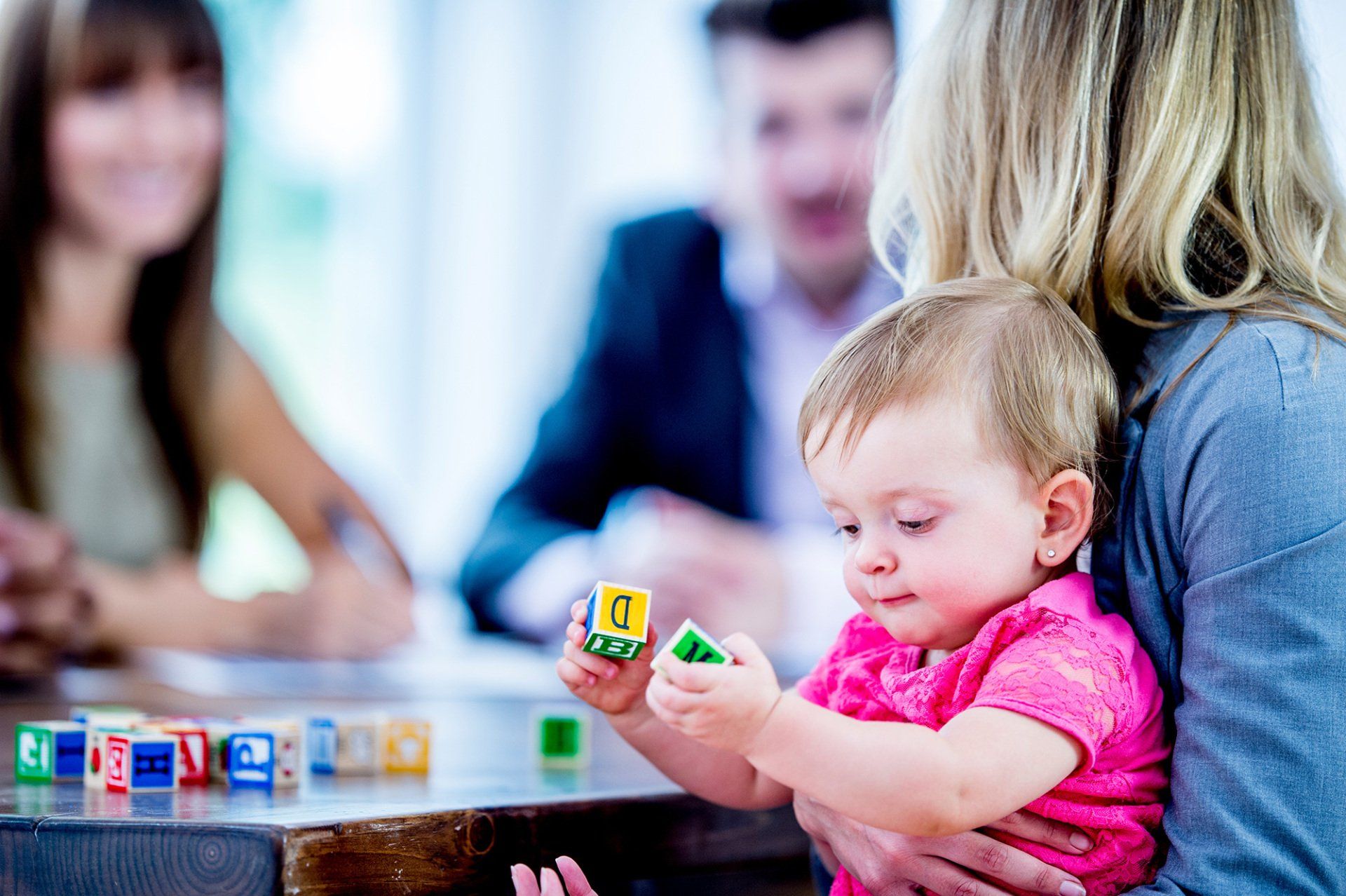 A mother is holding her baby daughter while attending an office meeting. The baby is smiling while playing with colourful wooden blocks.
