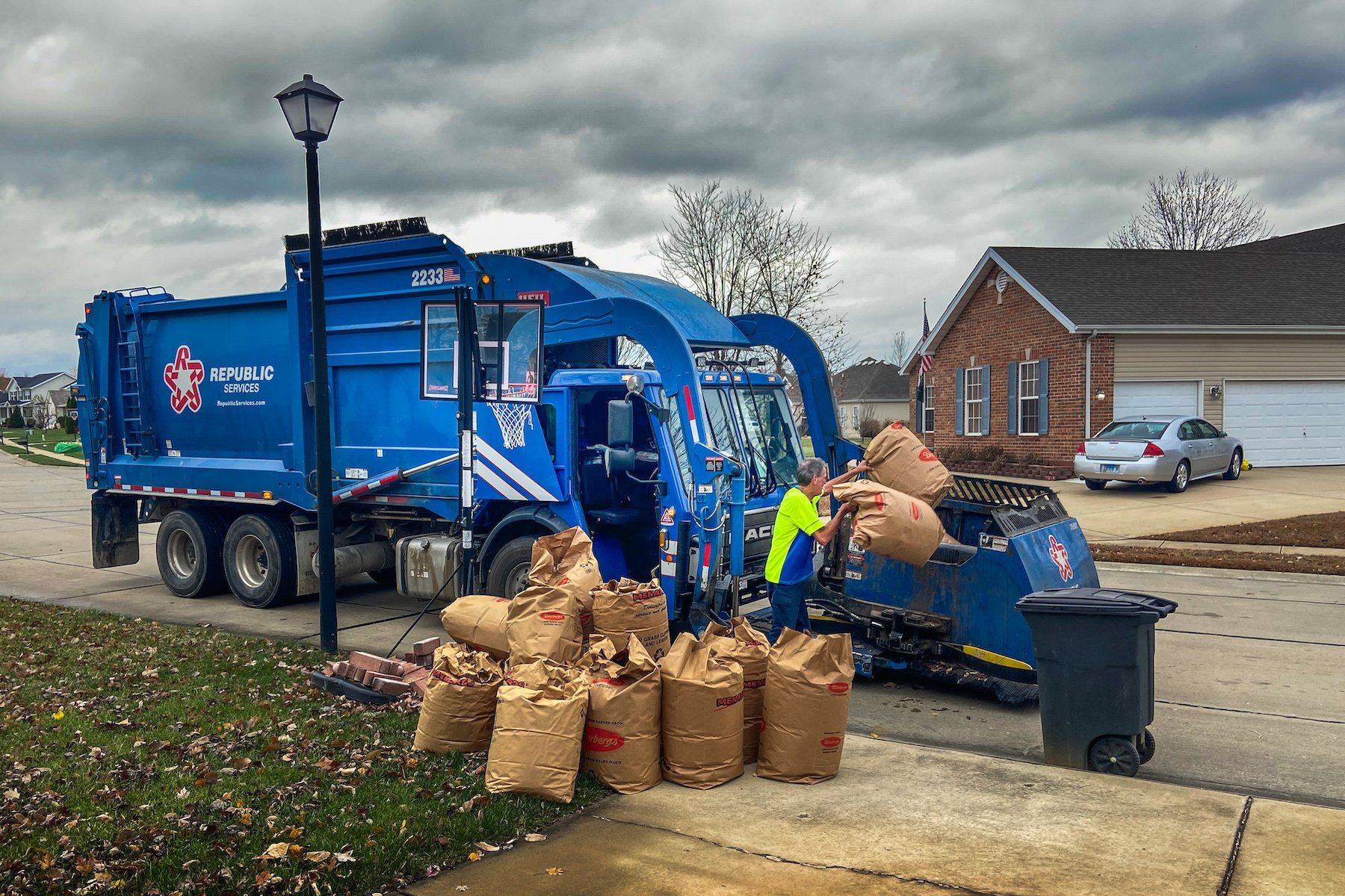 Waste hauler truck with driver collecting compostable yard waste in lawn and leaf bags from a residential area.