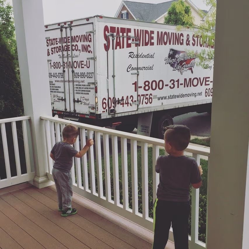 Two young children watch a State-Wide Moving truck from a porch.