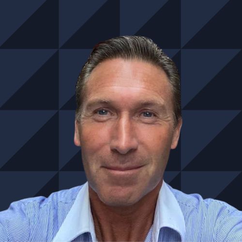 Man with short, slicked-back hair, wearing a blue collared shirt, smiling, set against a geometric blue background.