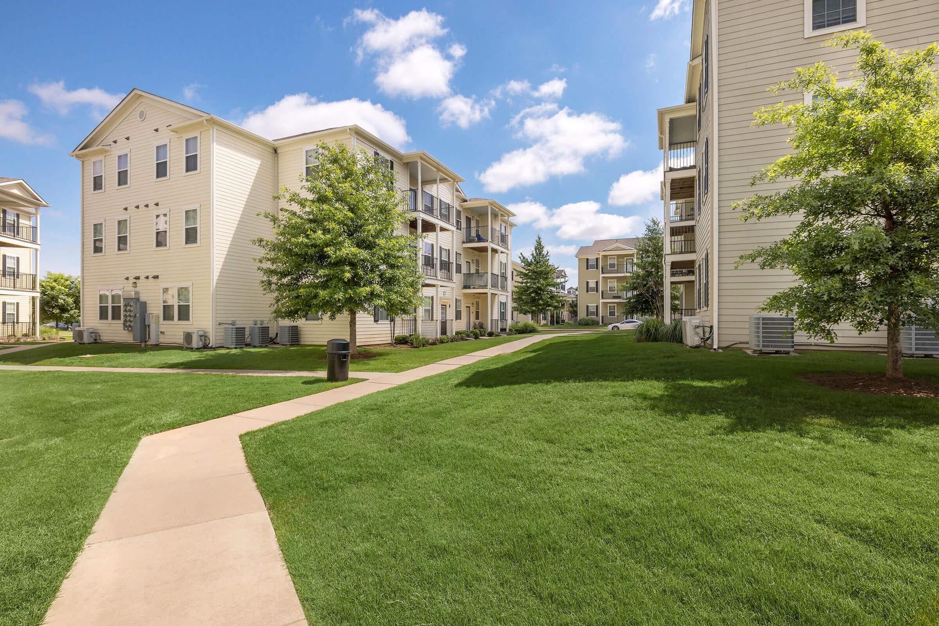 Apartment complex with beige buildings, green lawn, trees, and a walkway on a sunny day.