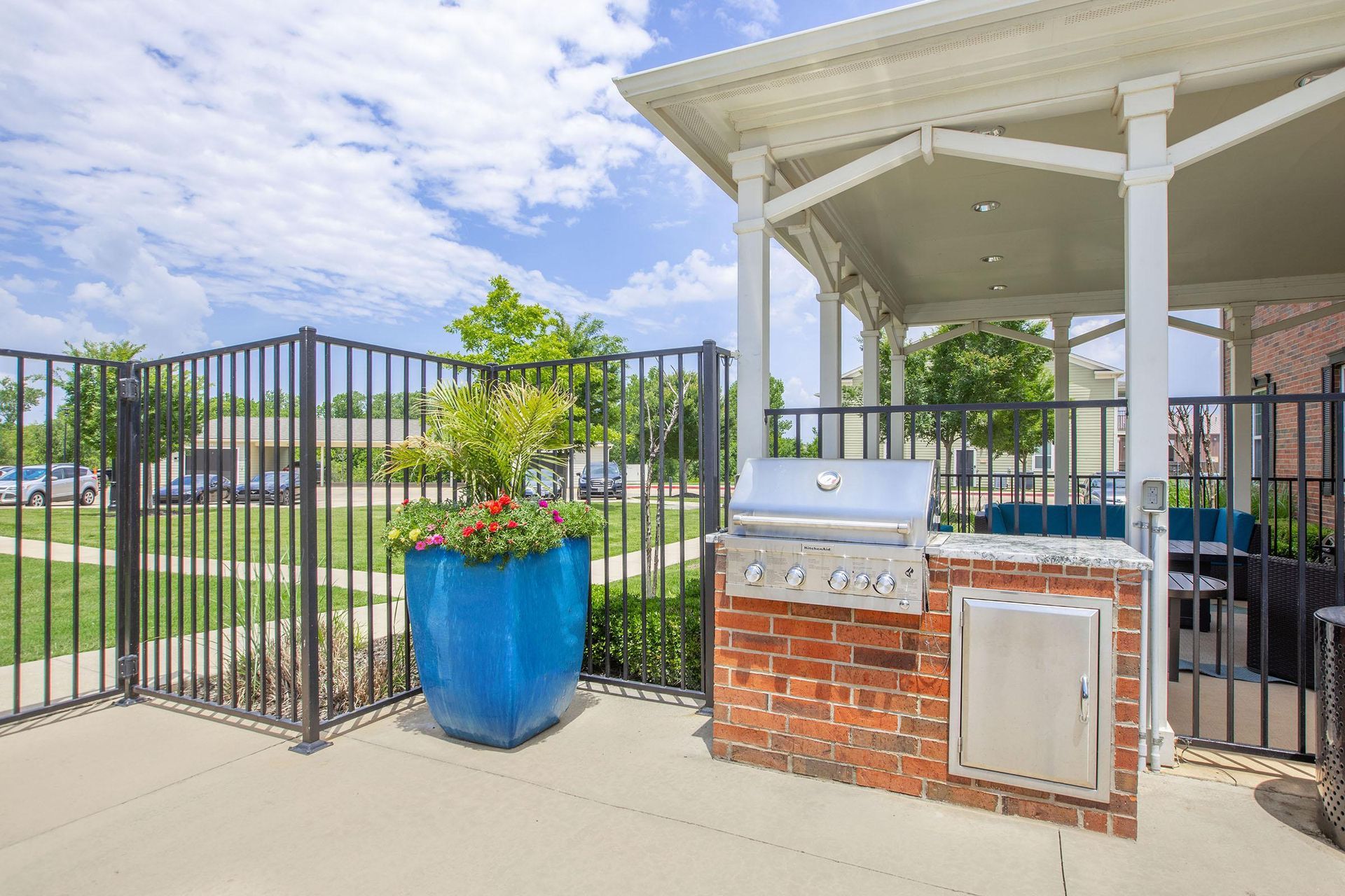 Outdoor grilling station with brick base, stainless steel grill, and blue flower pot near covered patio and black fence.