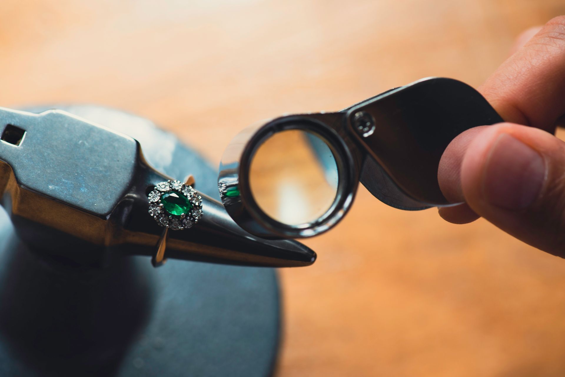 A hand examines a ring with a magnifying glass; the ring features a green gem.