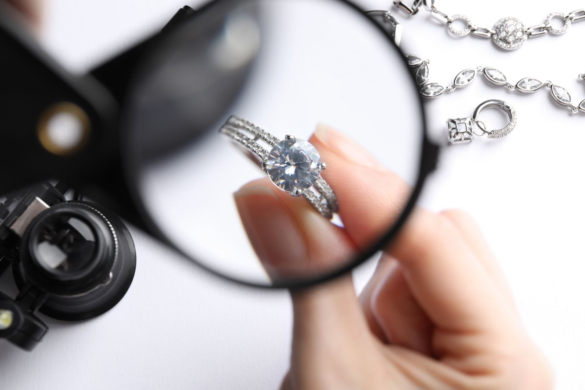 Person examining a diamond ring with a magnifying glass, with other jewelry in the background.