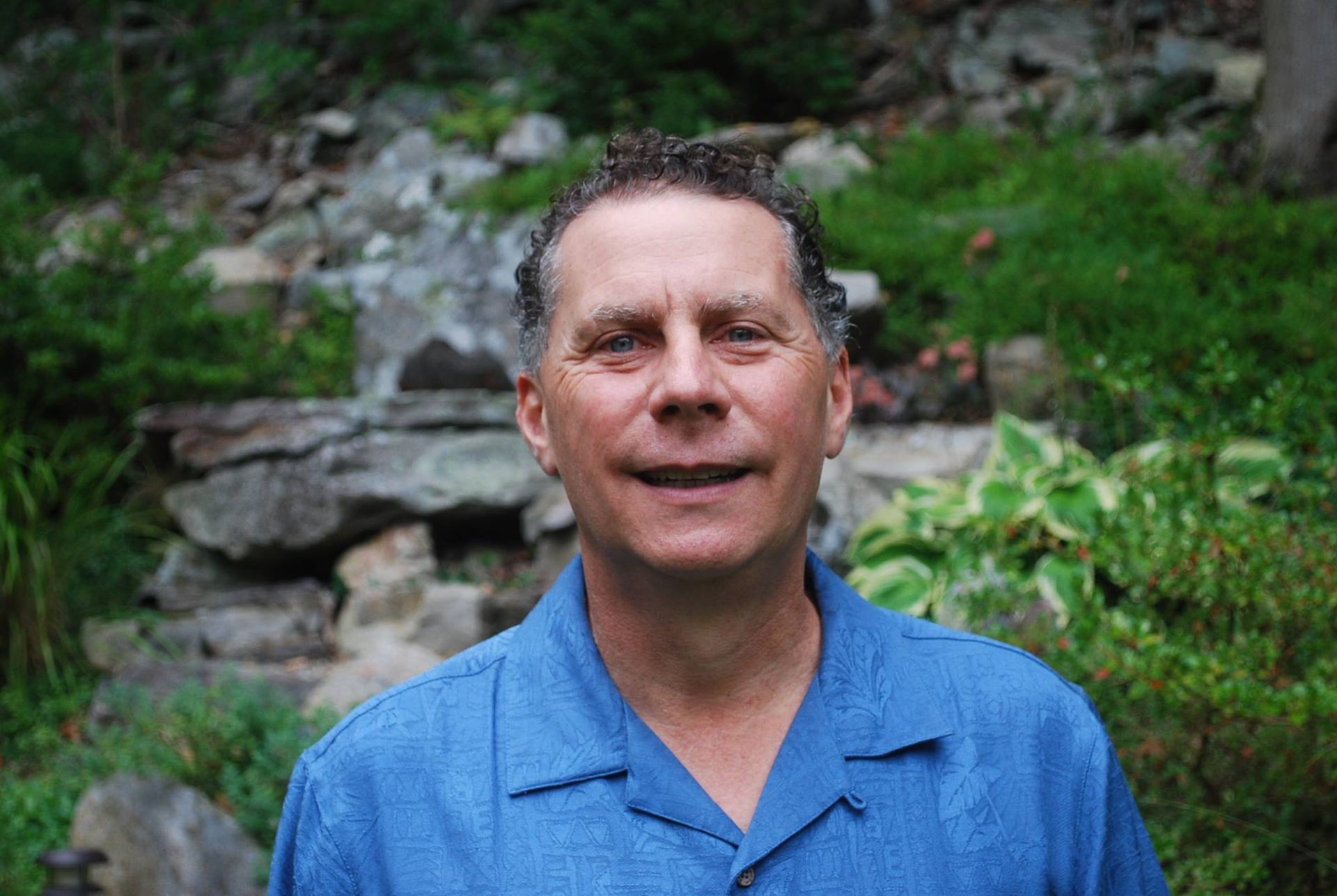 A man in a blue shirt is smiling in front of a waterfall.