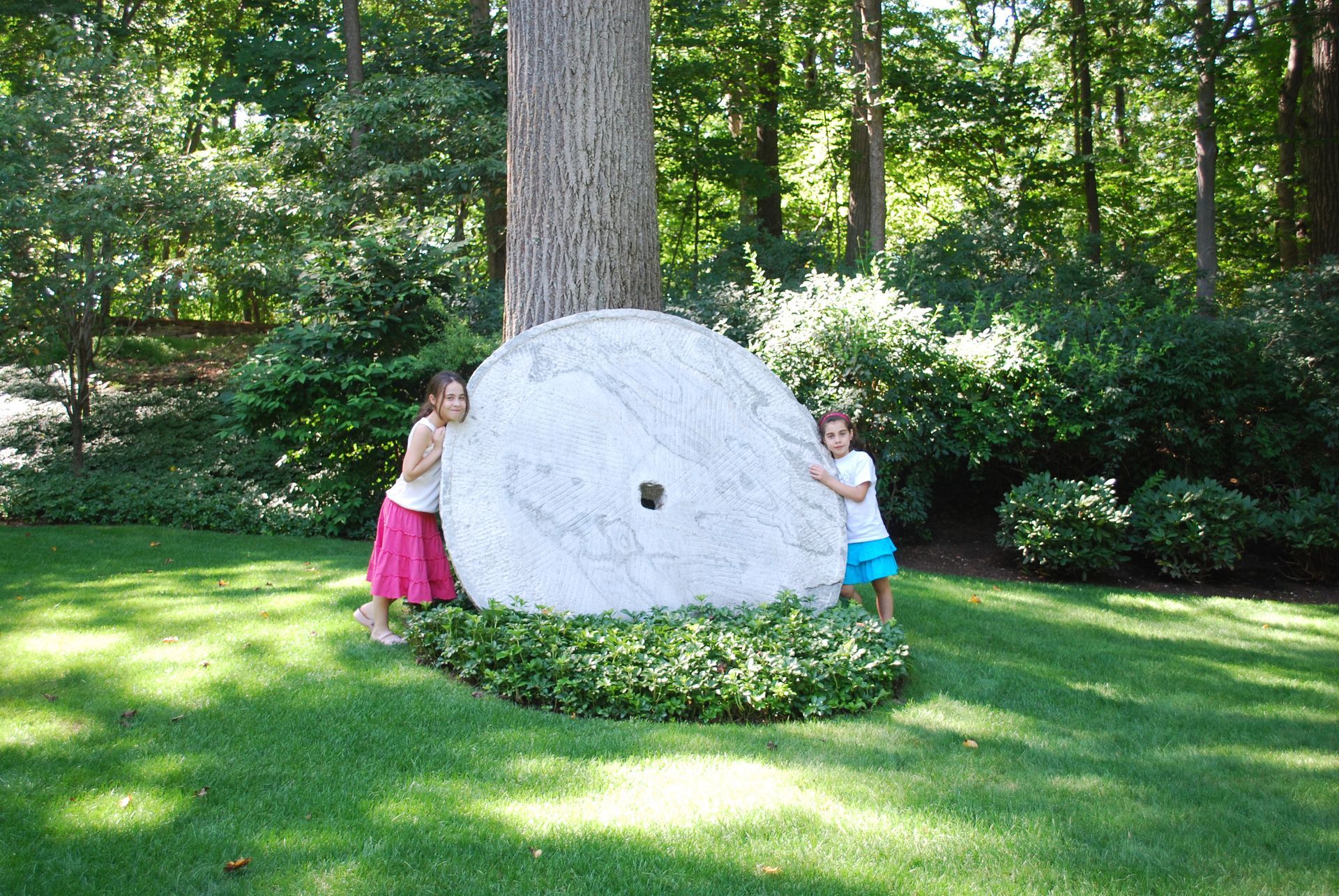 Two little girls are standing next to a large stone in the grass