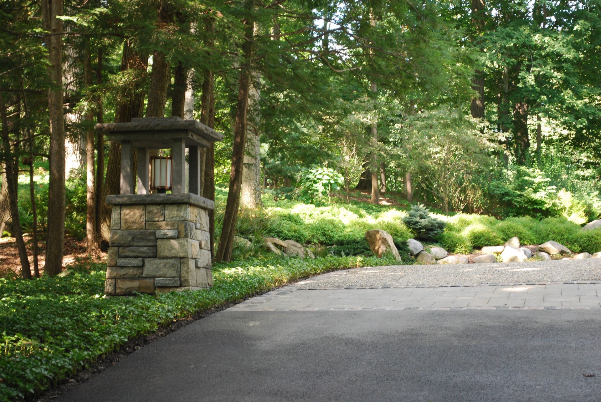 A stone pillar with a light on it is in the middle of a driveway surrounded by trees.