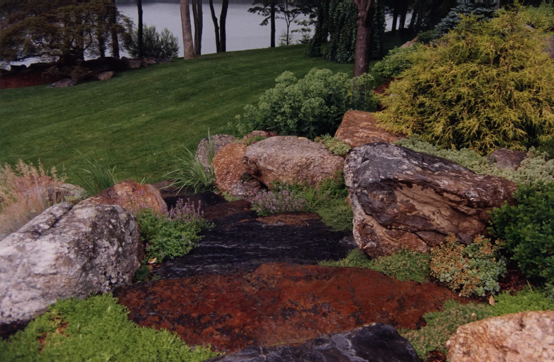 A lush green lawn surrounded by rocks and bushes with a lake in the background