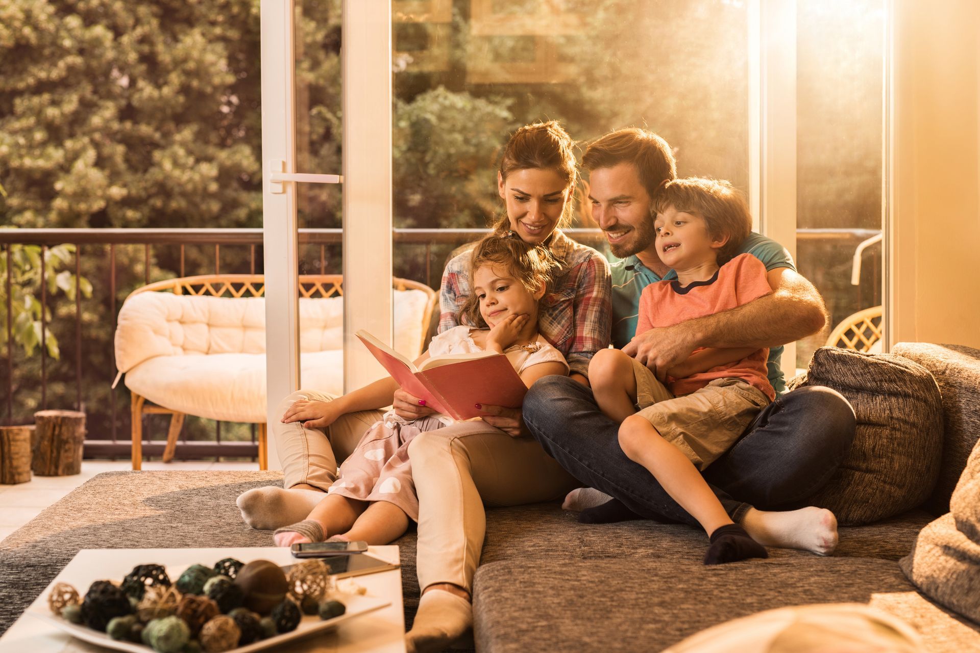 Family reading a book together on a couch, bathed in warm sunlight. Mother, father, daughter, and son sharing a story.