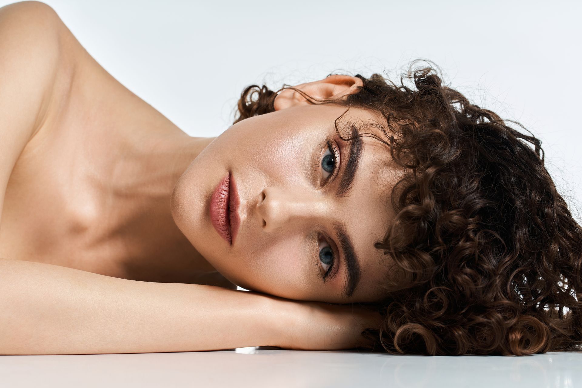 Woman with curly hair reclines, resting on arms, looking towards viewer.