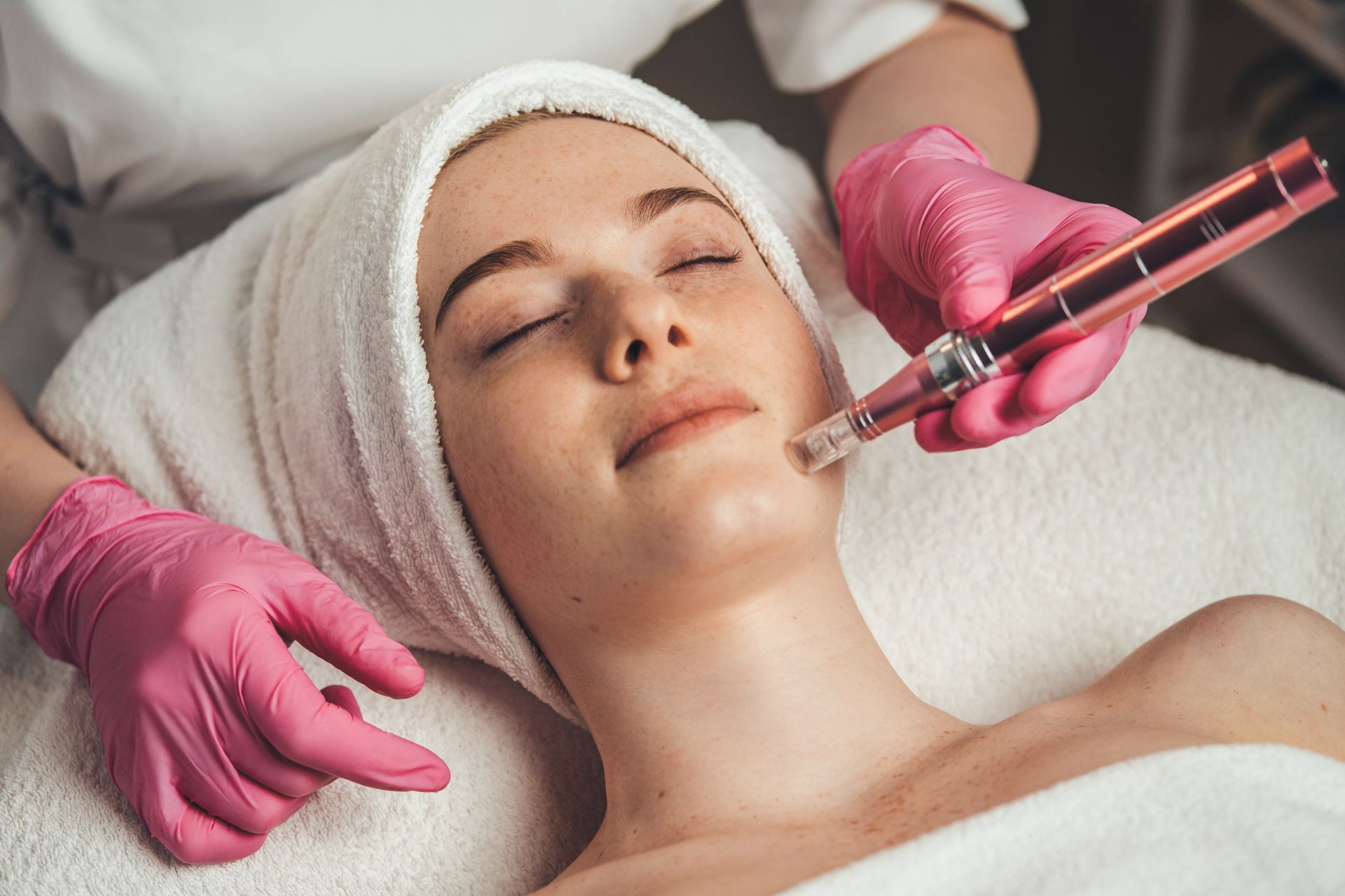 Woman receiving microneedling treatment on her face; hands in pink gloves; white towel on head.