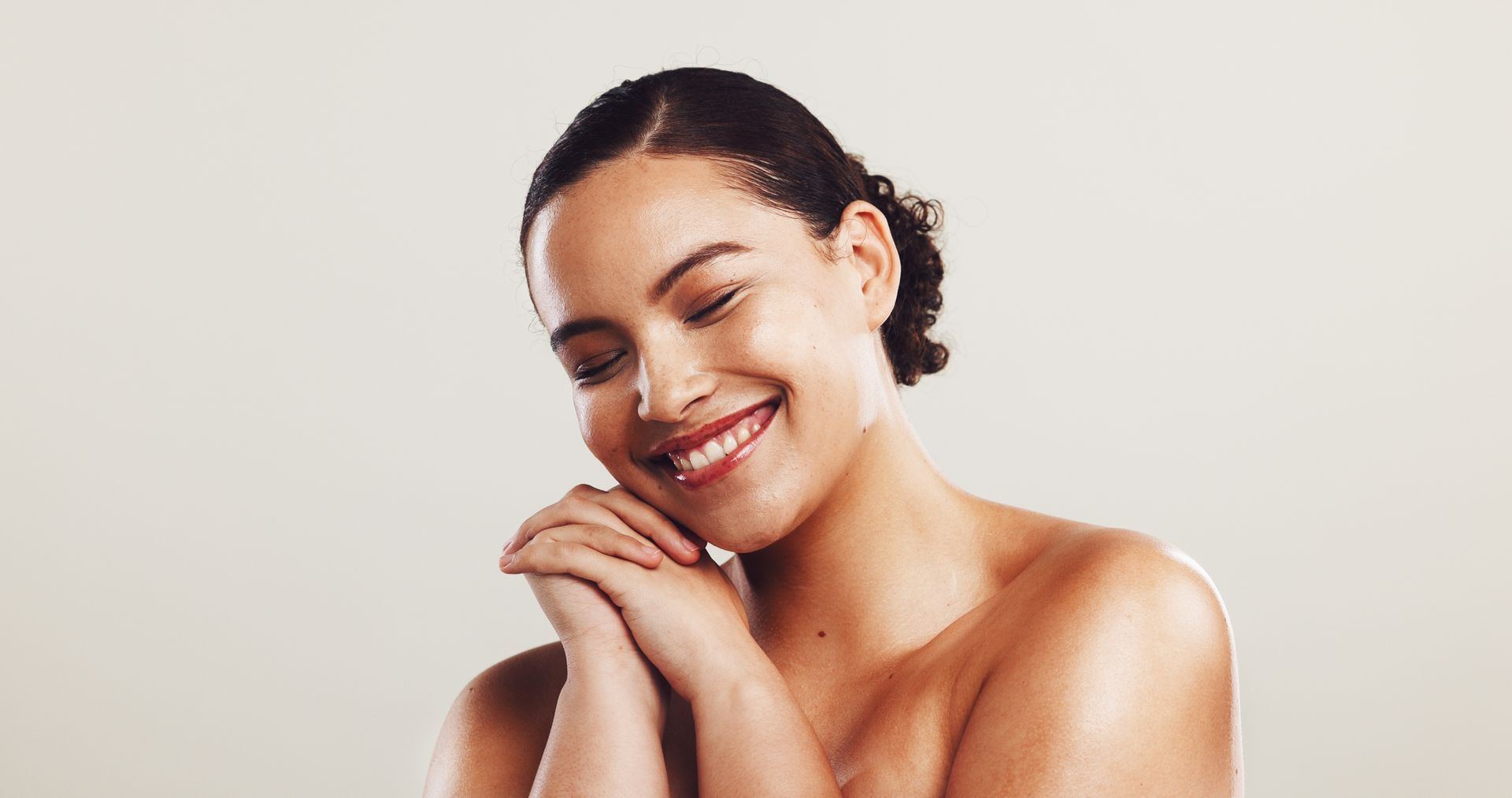 Smiling woman with bare shoulders resting her chin on clasped hands against a light background