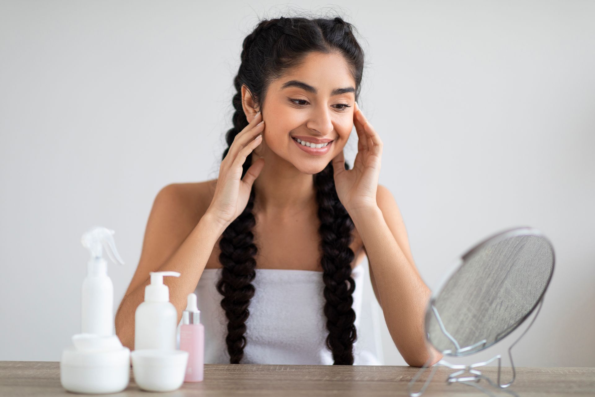 Woman applying skincare on her face beside bottles and a mirror in a bright bathroom