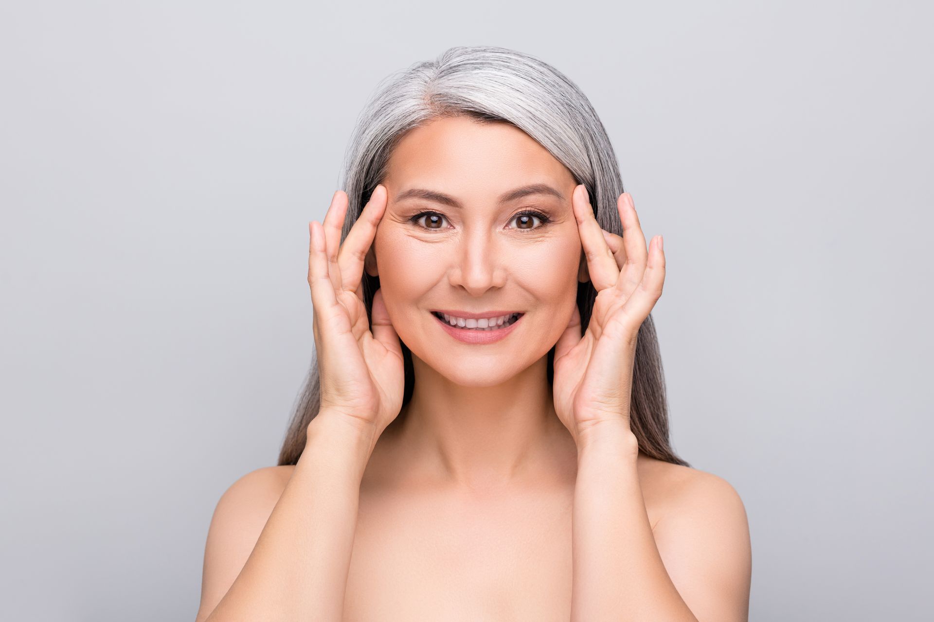 A woman with gray hair is smiling and touching her face.