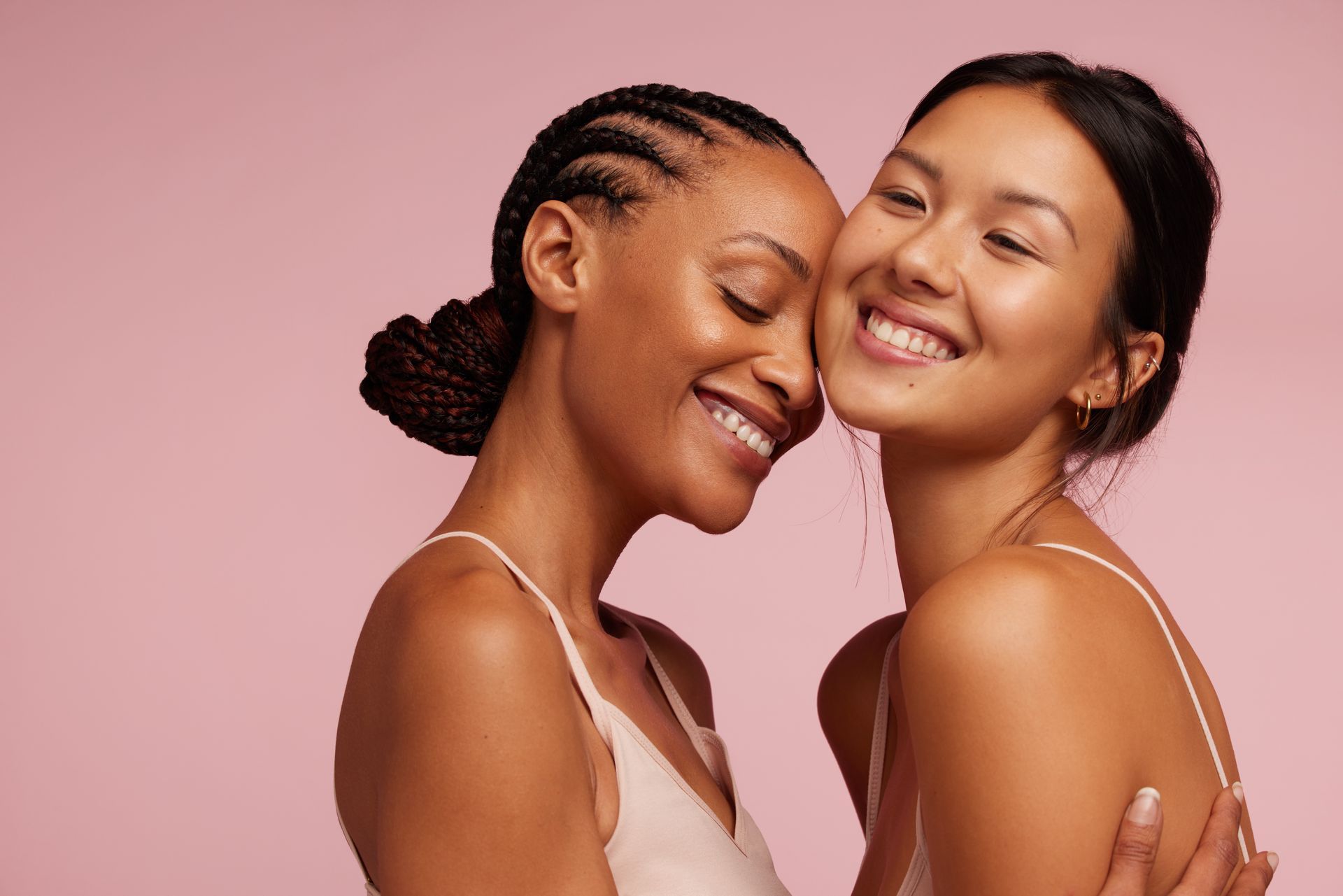 Two women are hugging each other on a pink background and smiling.