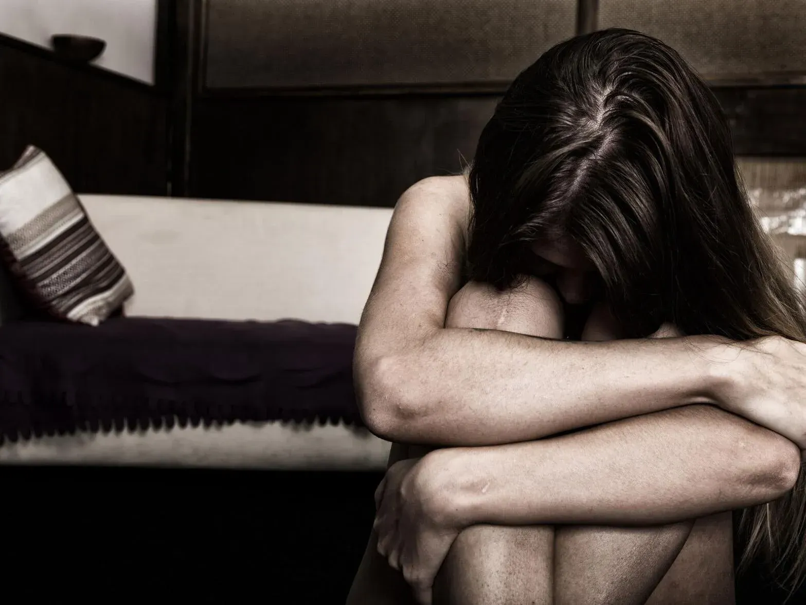 Person sitting curled up on a couch, head down and arms wrapped around knees, in a dark room.
