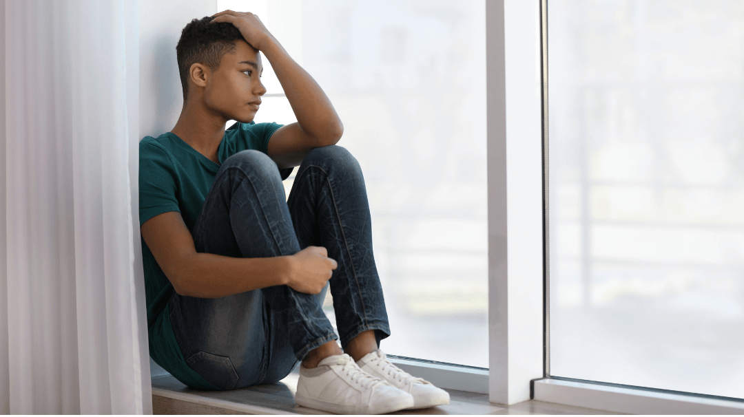 Person sitting on the floor by a window, knees hugged, looking out thoughtfully.