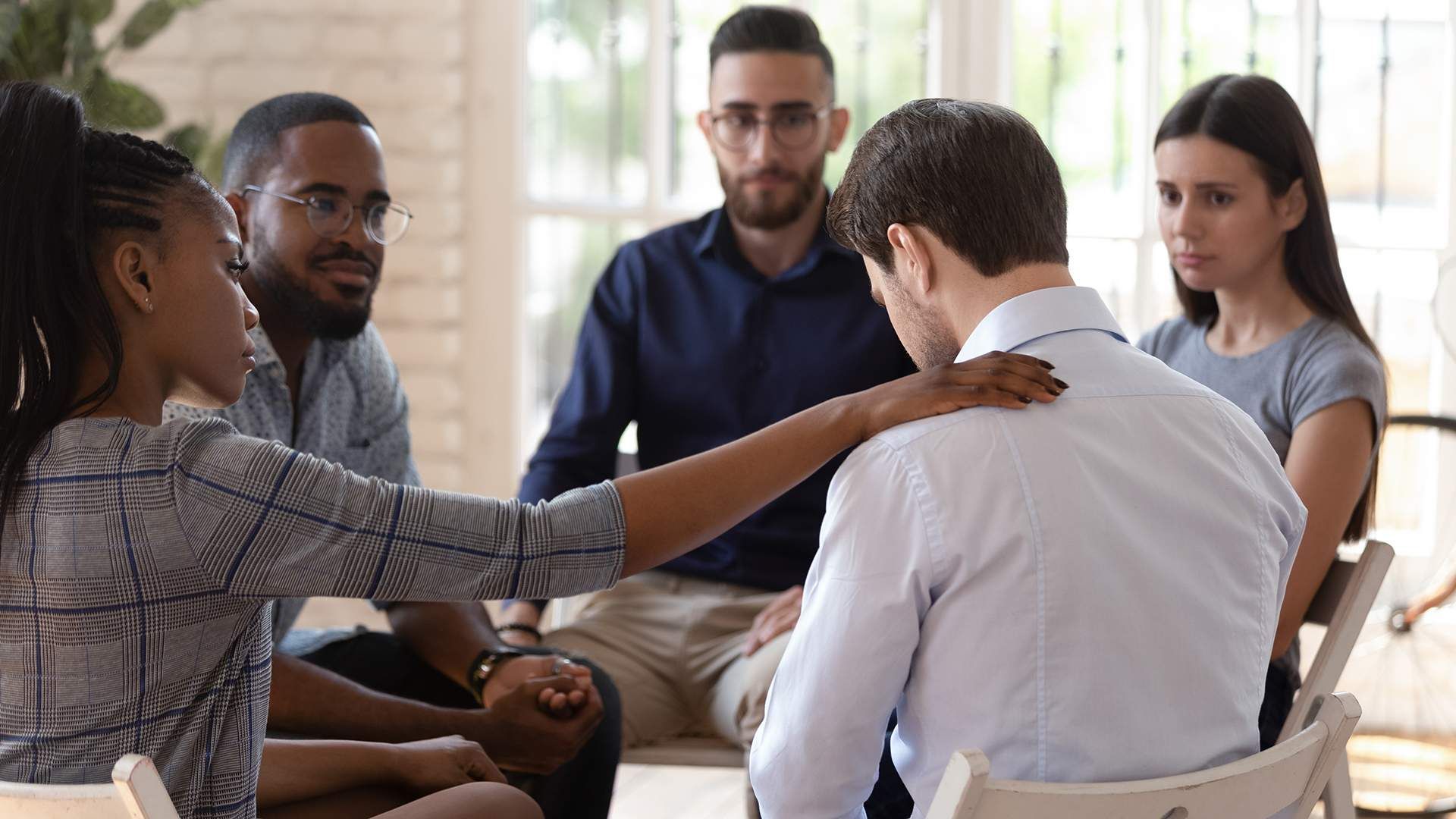 Man on sofa holds pillow, looks down, talking with therapist in office.