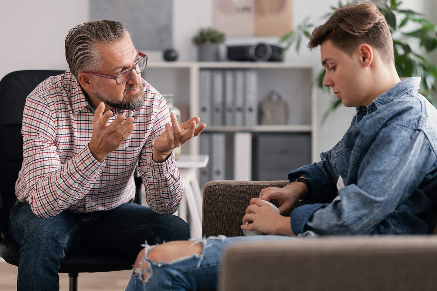 Man in glasses gestures while talking to a young person sitting on a couch. Office setting.