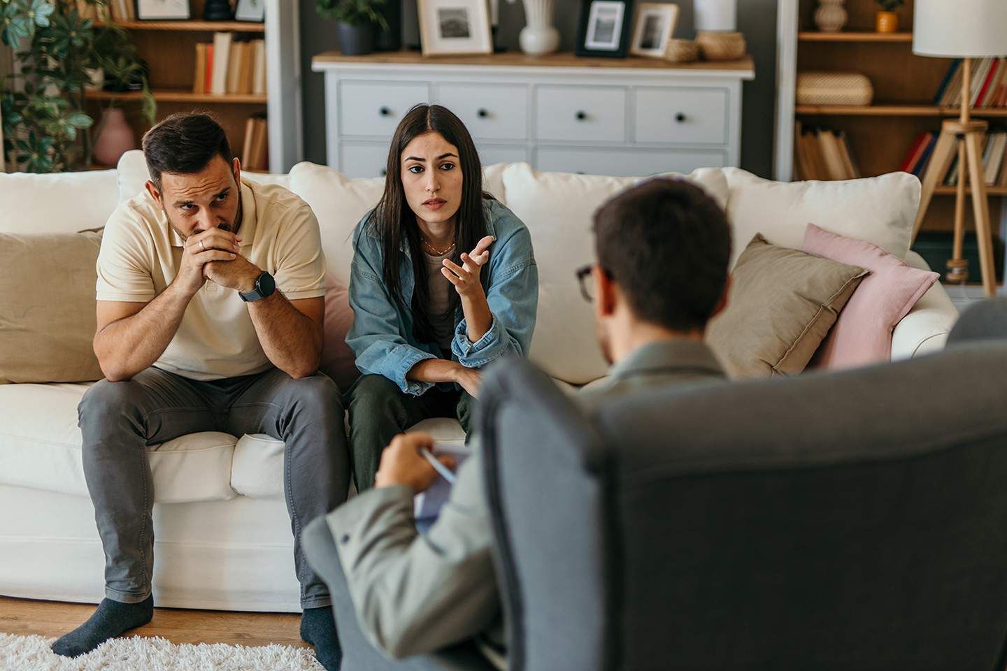 Couple in therapy, looking distressed, talking with a therapist seated in a chair.