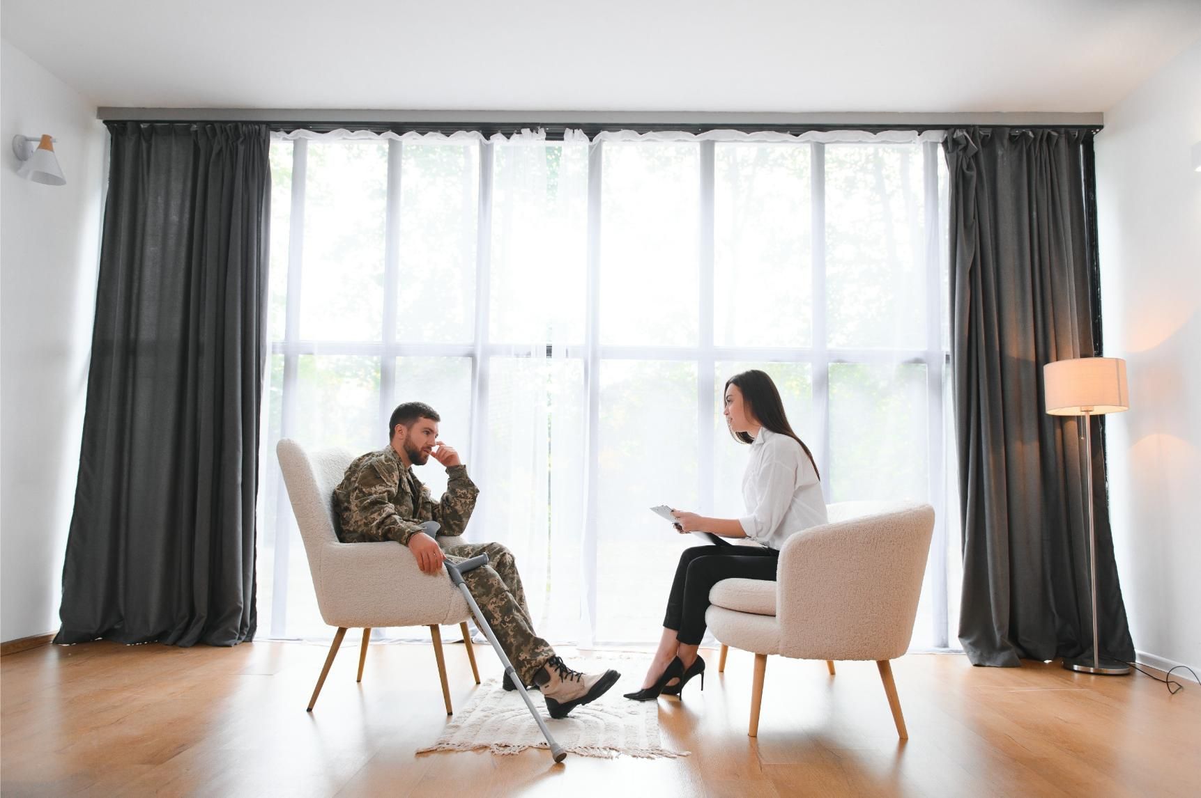 A soldier using a crutch speaks with a professional in a bright office with large windows and gray curtains.