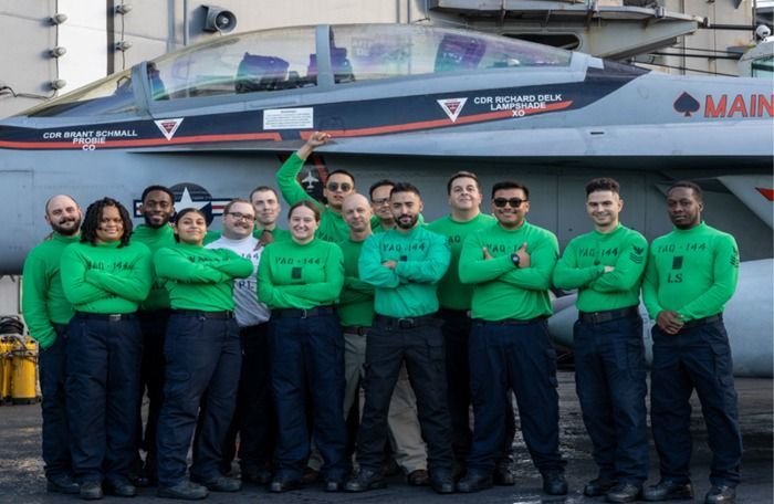 Navy crew in green shirts posing with arms crossed in front of a fighter jet on an aircraft carrier