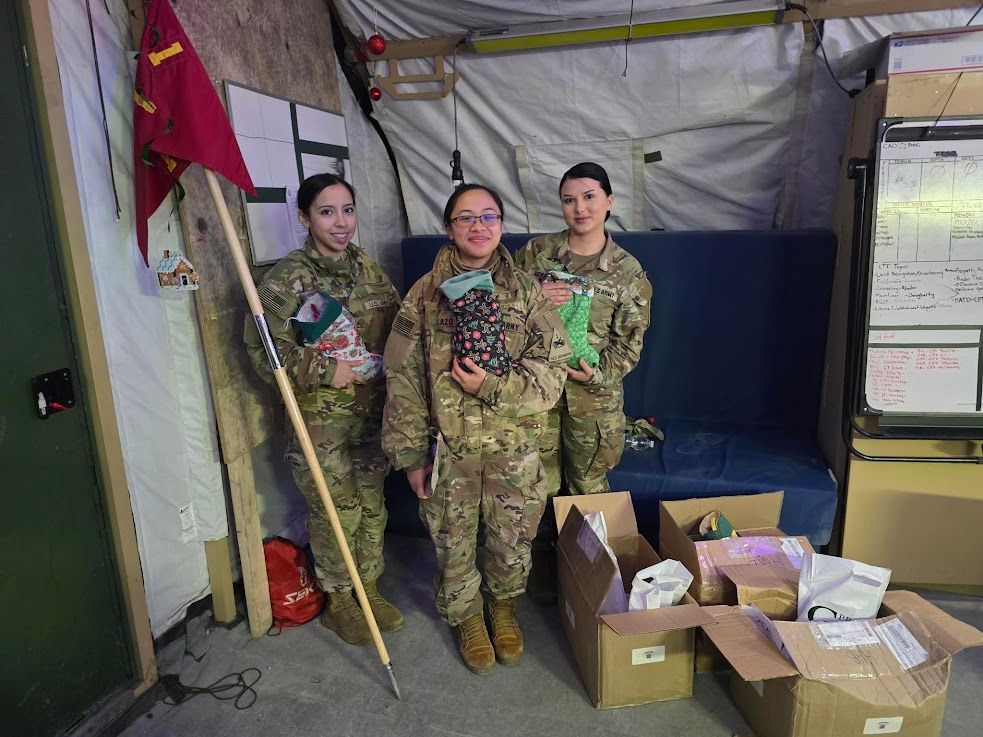 Three female soldiers, in uniform, hold gifts inside a tent