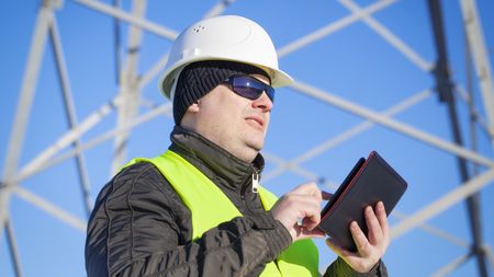 A person in a hard hat and high-visibility vest uses a tablet in front of a metal transmission tower against a blue sky.