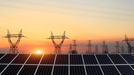 Solar panels in the foreground with high-voltage electricity transmission towers set against a sunset sky.