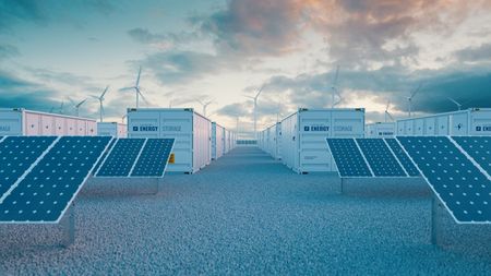 Rows of solar panels and shipping-container battery storage units under a cloudy sky with wind turbines in the distance.