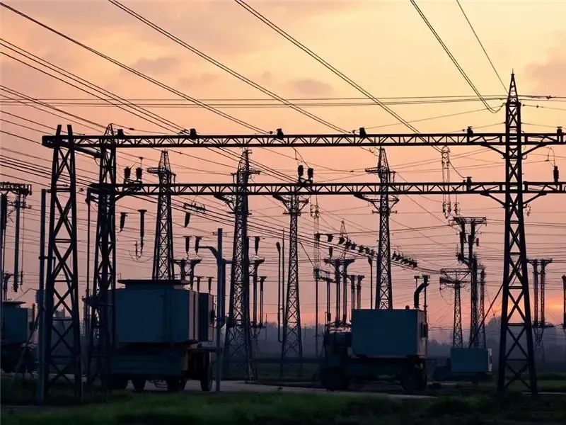 Two technicians in hard hats inspect electrical panel, one using a laptop in a control room.