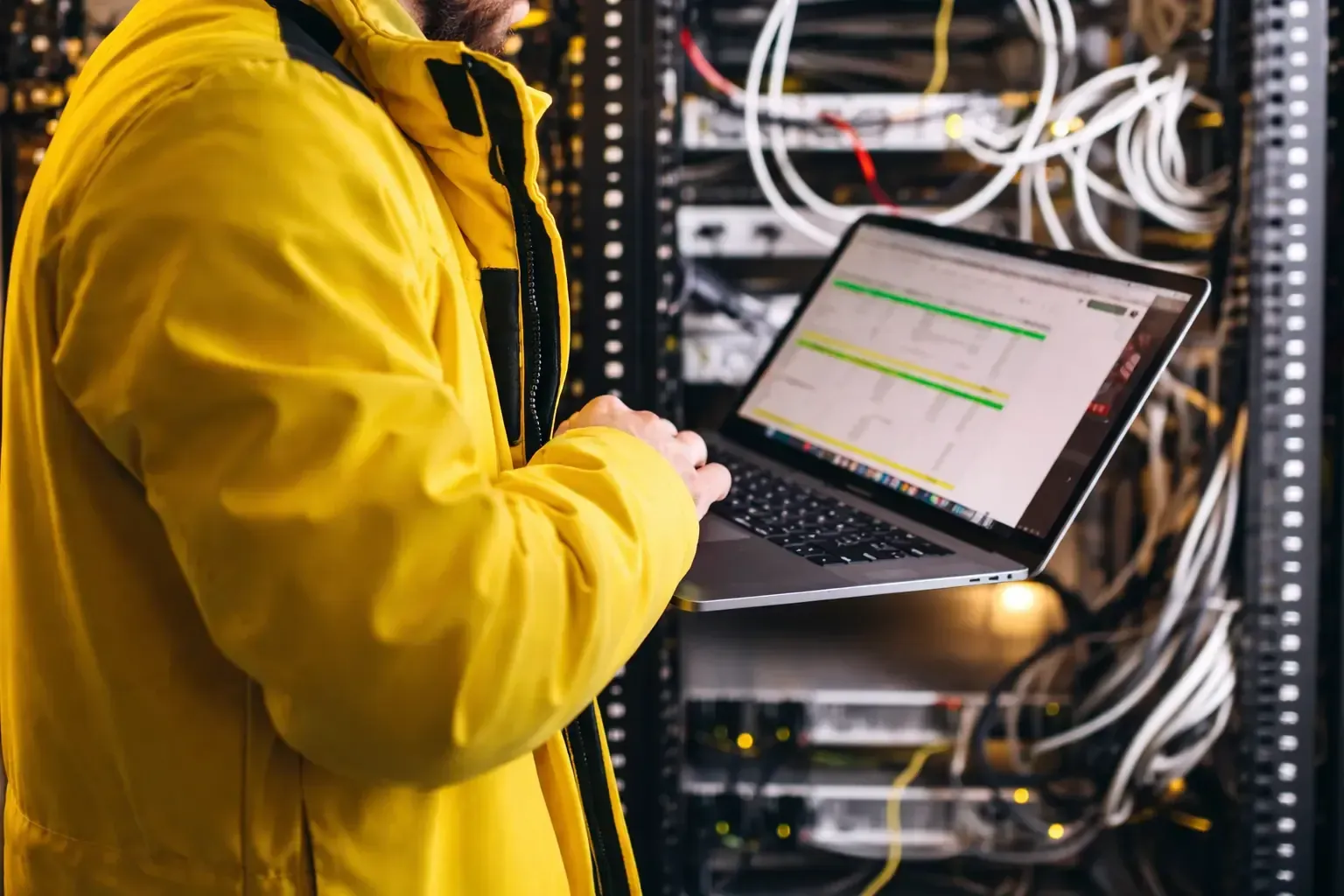 IT technician in a yellow jacket using a laptop to monitor servers in a data center with organized cables and systems
